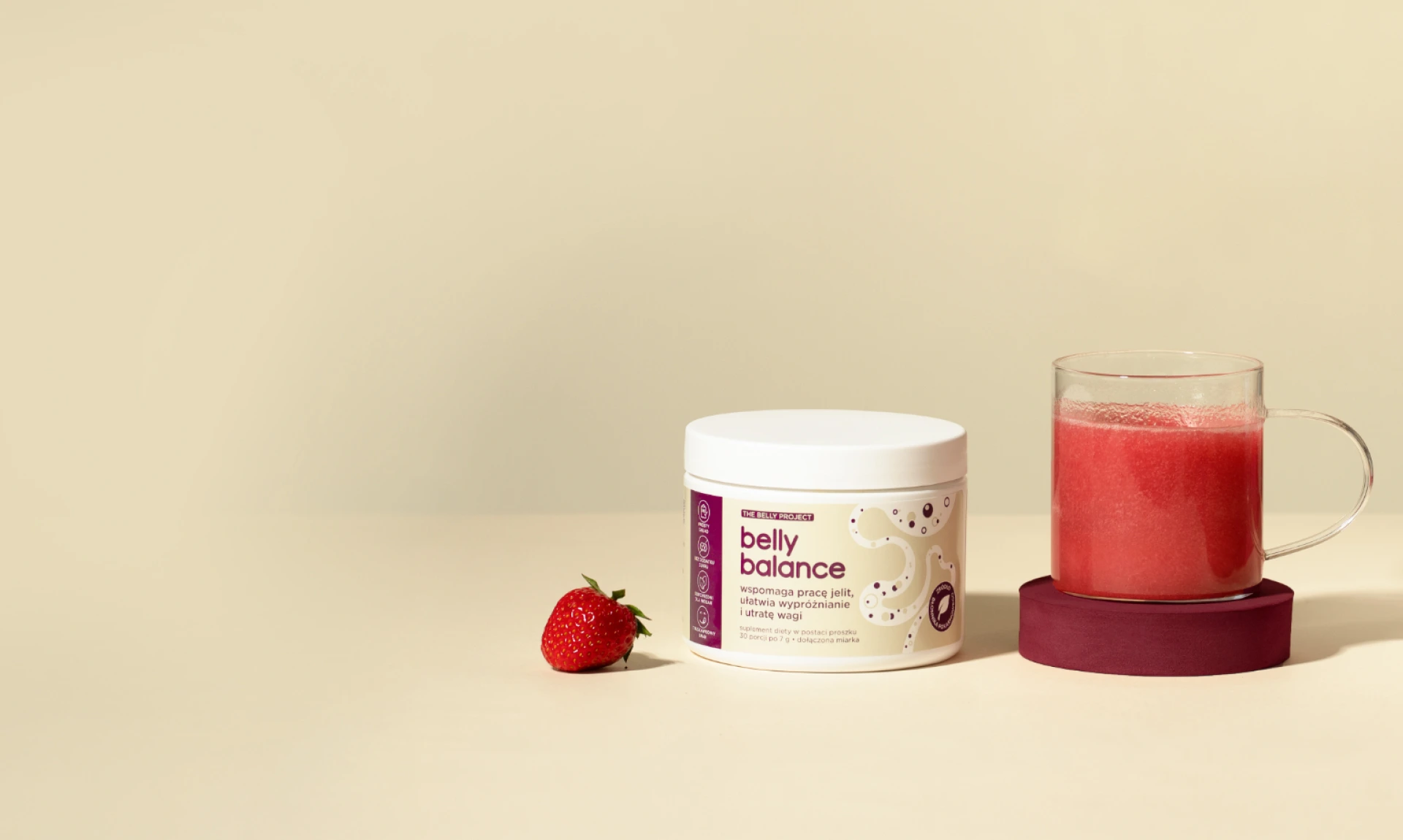Opakowanie suplementu diety BellyBalance z białą pokrywką obok szklanego kubka wypełnionego przygotowanym suplementem, ustawionego na drewnianej podstawce, z świeżą truskawką na beżowym tle.