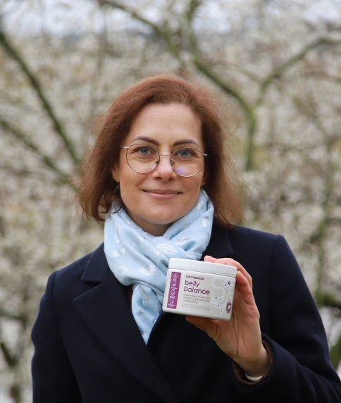 Kobieta z rudobrązowymi włosami w okularach, ubrana w ciemny płaszcz i jasnoniebieski szalik, trzymająca opakowanie suplementu diety BellyBalance, stojąca na zewnątrz z drzewami w tle.