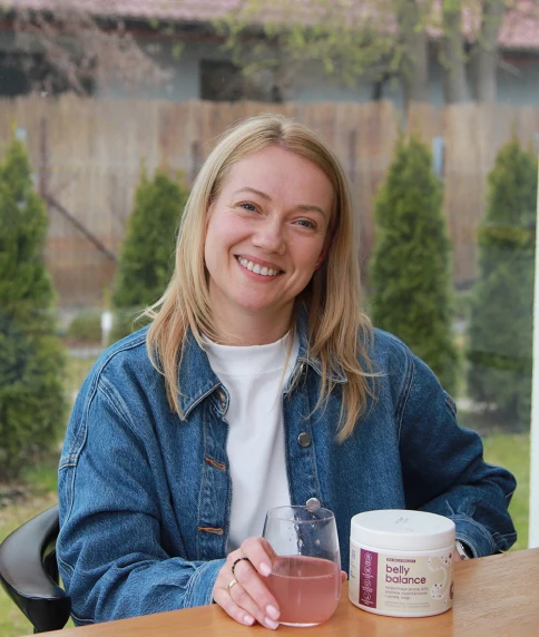 Uśmiechnięta blond kobieta ubrana w jeansową kurtkę i biały t-shirt, siedząca przy stoliku na zewnątrz ze szklanką czerwonego napoju i opakowaniem suplementu diety BellyBalance, z drewnianym płotem i zielonymi roślinami w tle.