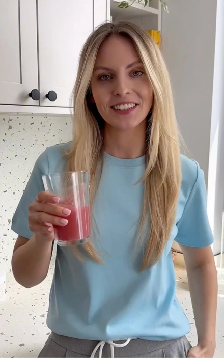Uśmiechnięta blond kobieta ubrana w jasnoniebieską koszulkę, trzymająca szklankę różowego smoothie w jasnej białej kuchni z białymi szafkami i nakrapianym blatem.